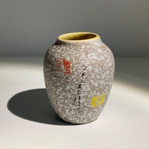 Könnte beinhalten: Eine kleine, gesprenkelte Keramikvase mit runder Form. Die Vase hat ein hellbraunes und weiß gesprenkeltes Muster mit einem gelben Innenrand. Sie hat rote und gelbe Akzente und ein schwarzes Liniendesign. Die Vase wird wahrscheinlich zu dekorativen Zwecken verwendet.
