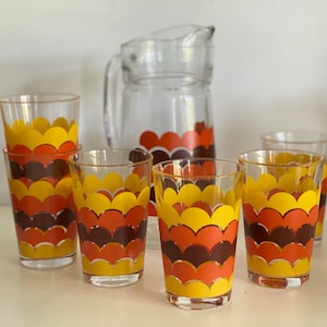 Può includere: Un set di vetreria vintage con una caraffa in vetro trasparente e sei bicchieri coordinati. I bicchieri e la caraffa sono decorati con un motivo ripetuto di forme a conchiglia gialle, arancioni e marroni. Il set è probabilmente della metà del secolo.