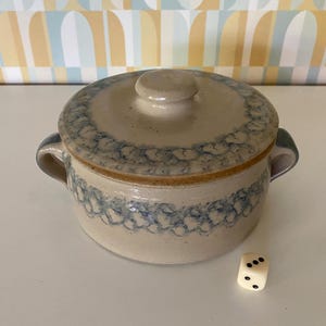 Può includere: Una zuppiera in ceramica con coperchio e manici, con base beige e motivo floreale blu sul bordo e sul corpo. Un dado bianco con punti neri è accanto alla ciotola.