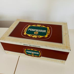 Può includere: Una scatola rettangolare rossa e bianca con un logo dorato e verde che dice "Pompadour" e il testo "Inhalt 500 Teebeutel".