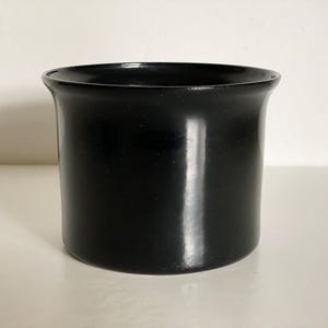 Könnte beinhalten: Ein schwarzer zylindrischer Keramikbehälter mit leicht ausgestelltem Rand. Der Behälter hat eine glatte, glänzende Oberfläche. Er steht vor einem weißen Hintergrund. Das Design ist schlicht und modern, für verschiedene Zwecke geeignet.
