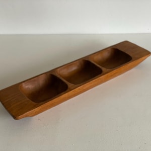 Op de afbeelding: Een lange, rechthoekige houten serveerschaal met drie verzonken compartimenten. De schaal is gemaakt van lichtbruin hout en heeft een gladde, gepolijste afwerking. De compartimenten zijn gelijkmatig verdeeld en zijn bedoeld voor het serveren van snacks of hapjes.