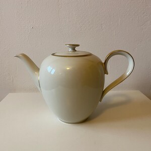 May include: A white porcelain teapot with a gold trim around the lid and spout. The teapot has a rounded body and a curved handle.