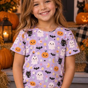 Könnte beinhalten: Ein kurzärmeliges T-Shirt in Helllila mit einem durchgehenden Druck von Halloween-Motiven, darunter orangefarbene Kürbisse, weiße Geister, schwarze Katzen, Fledermäuse, gelbe Sterne und Mondsicheln auf einem Rundhalsausschnitt.