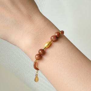 Può includere: Un braccialetto con perline rotonde in pietra del sole marrone scintillante, alternate a piccoli distanziatori dorati e una singola perla centrale di forma ovale color oro. Il braccialetto include una delicata catenella di estensione con un piccolo ciondolo.