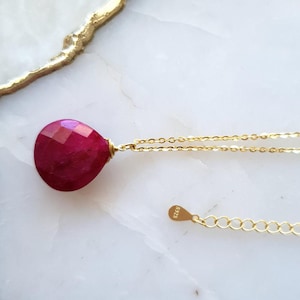 May include: A gold chain necklace with a teardrop-shaped ruby pendant. The pendant is a deep red color and has a faceted surface.