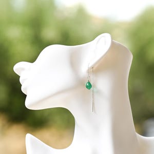 Może przedstawiać: Para srebrnych kolczyków z pojedynczym zielonym kamieniem szlachetnym zwisającym z każdego łańcuszka.