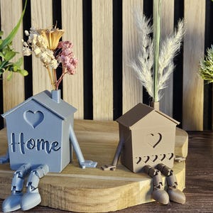 Könnte beinhalten: Eine dekorative Wohnkultur-Anordnung mit zwei hausförmigen Vasen mit Herz-Ausschnitten und dem Wort "Home". Die Vasen haben Beine und Schuhe und stehen auf einer Holzplattform. Die Anordnung umfasst künstliche Blumen und Pflanzen.