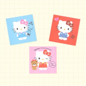 May include: Three square stickers featuring Hello Kitty. The blue sticker shows Hello Kitty with a milk bottle and the text "VERY DELICIOUS". The red sticker shows Hello Kitty with a heart and "HELLO KITTY". The pink sticker shows Hello Kitty and a bear with "Together in fun forever!" and "Stuck with me!".