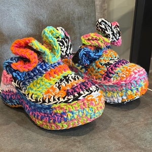 May include: A pair of handmade crochet slippers featuring a vibrant, multi-colored yarn pattern in shades of neon pink, orange, blue, yellow, and black. Each slipper has a thick, textured sole and a ruffled, decorative accent on the top front.