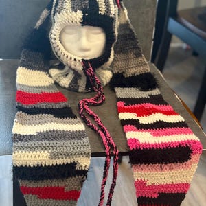 May include: A handmade crochet hat with long ear flaps. The hat features a striped pattern in black, white, gray, red, and pink. The ear flaps have a striped pattern and are connected with a pink and black braided cord.