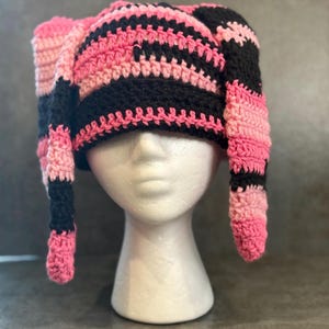 May include: A handmade crochet hat in pink and black stripes. The hat has long, ear-like extensions. The hat is displayed on a white mannequin head against a gray background. The hat is a unique, colorful accessory.