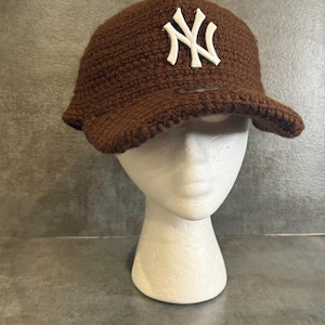 May include: A brown crocheted baseball cap with a white "NY" logo on the front. The cap has a curved brim and is displayed on a white mannequin head. The background is a textured gray surface.
