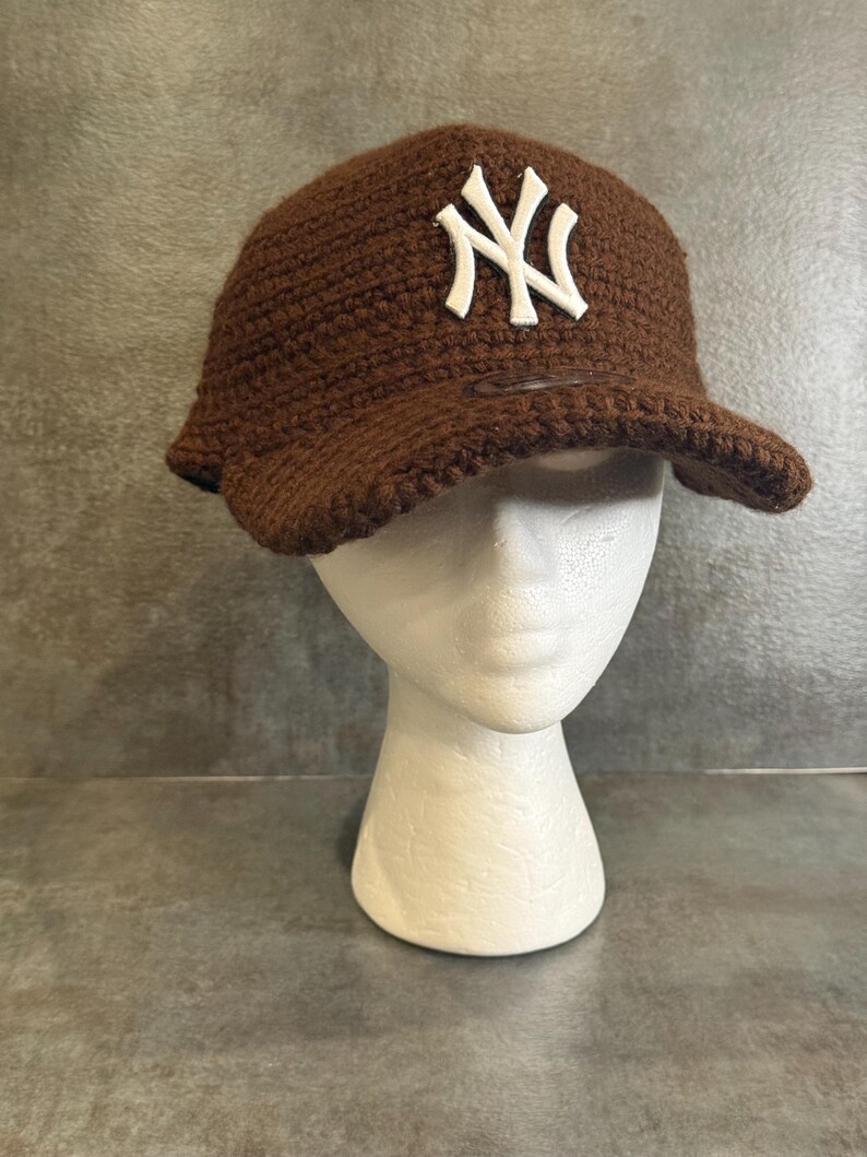 May include: A brown crocheted baseball cap with a white "NY" logo on the front. The cap has a curved brim and is displayed on a white mannequin head. The background is a textured gray surface.