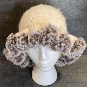 May include: Cream-colored knitted hat with a fluffy, faux fur trim in shades of brown and gray. The hat is displayed on a white mannequin head, showcasing its shape and texture. A cozy winter accessory.