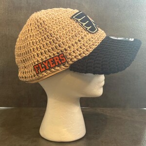 May include: A tan crocheted hat with a black brim. The hat features two patches with the word "FLYERS" in orange letters and a logo. The hat is displayed on a white mannequin head.