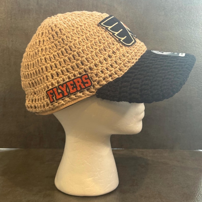 May include: A tan crocheted hat with a black brim. The hat features two patches with the word "FLYERS" in orange letters and a logo. The hat is displayed on a white mannequin head.