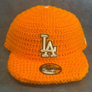 May include: Bright orange crochet baseball cap featuring the letters "LA" in white and tan. The cap has a curved brim and a "9FIFTY SNAPBACK" label. The hat is a vibrant accessory.