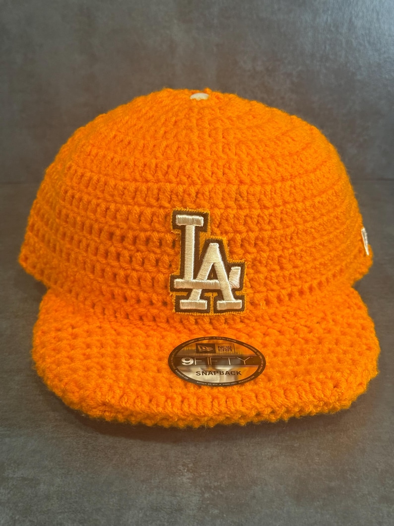 May include: Bright orange crochet baseball cap featuring the letters "LA" in white and tan. The cap has a curved brim and a "9FIFTY SNAPBACK" label. The hat is a vibrant accessory.