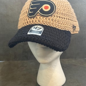 May include: A tan and black crochet hat with the Philadelphia Flyers logo patch. The hat has a black brim and a tag that reads "47 MVP Adjustable". The hat is displayed on a white mannequin head.
