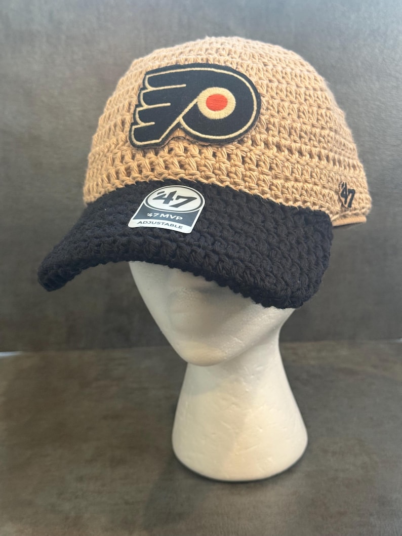 May include: A tan and black crochet hat with the Philadelphia Flyers logo patch. The hat has a black brim and a tag that reads "47 MVP Adjustable". The hat is displayed on a white mannequin head.
