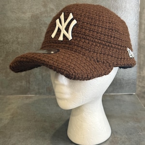 May include: A brown crocheted baseball cap with the New York Yankees logo in white. The cap has a curved brim and a New Era logo on the side. The hat is displayed on a white mannequin head.