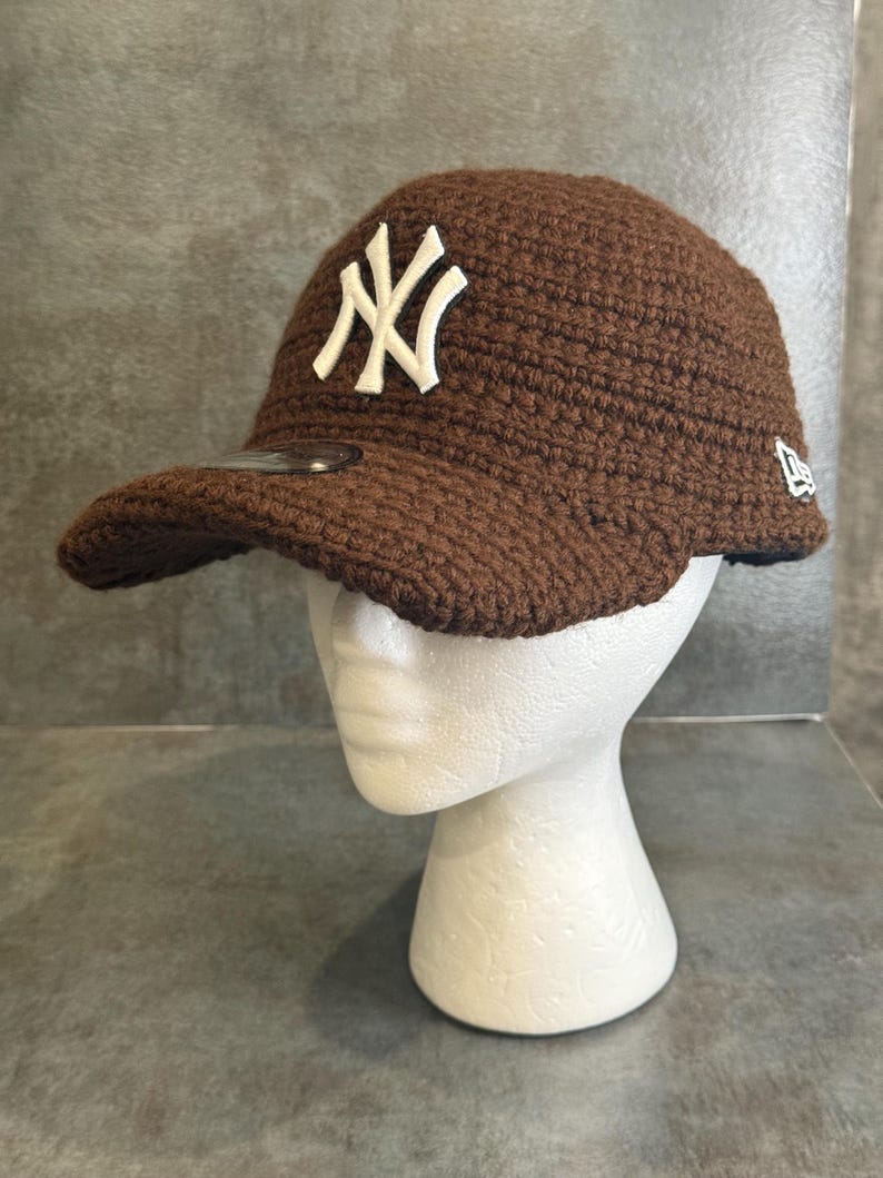 May include: A brown crocheted baseball cap with the New York Yankees logo in white. The cap has a curved brim and a New Era logo on the side. The hat is displayed on a white mannequin head.