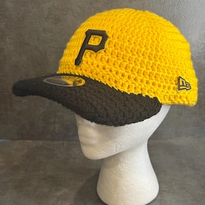 May include: A yellow and black crochet baseball cap with the letter "P" on the front. The cap has a black brim and the New Era logo on the side. The cap is displayed on a white mannequin head.