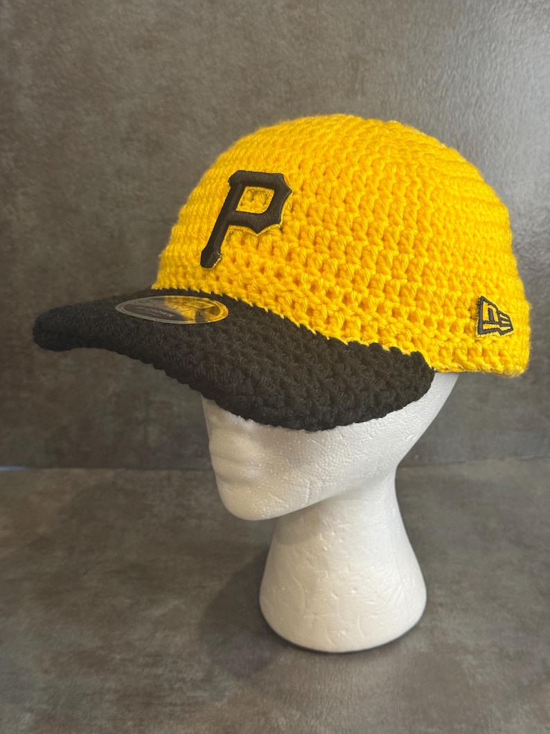 May include: A yellow and black crochet baseball cap with the letter "P" on the front. The cap has a black brim and the New Era logo on the side. The cap is displayed on a white mannequin head.