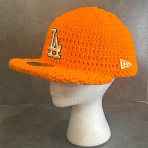May include: Bright orange crochet baseball cap with a curved brim. The cap features the letters "LA" in a cream color on the front and a white "New Era" logo on the side. The cap is displayed on a white mannequin head.
