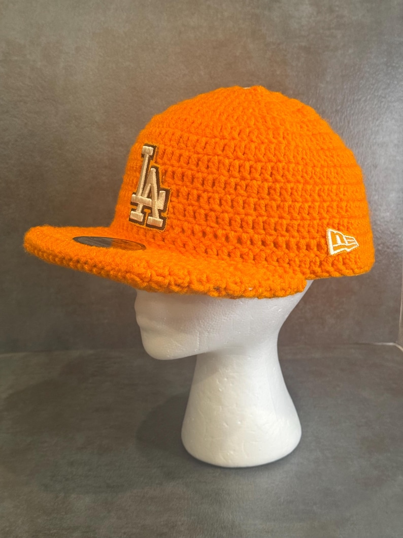 May include: Bright orange crochet baseball cap with a curved brim. The cap features the letters "LA" in a cream color on the front and a white "New Era" logo on the side. The cap is displayed on a white mannequin head.