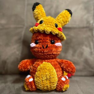 May include: A handmade orange and yellow crochet plush toy. The plush features a character wearing a yellow hat with black-tipped ears. The toy has black eyes, pink cheeks, and a yellow belly. The plush is sitting on a brown surface.