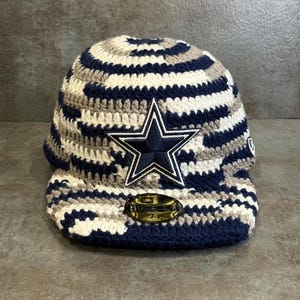 May include: A crocheted baseball cap in navy blue, white, and gray. The cap features a navy blue star logo with a white outline. The brim is navy blue, and the cap has a structured design.