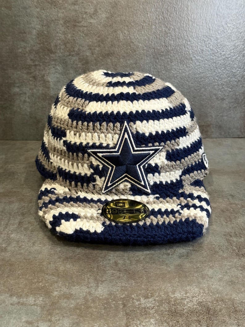 May include: A crocheted baseball cap in navy blue, white, and gray. The cap features a navy blue star logo with a white outline. The brim is navy blue, and the cap has a structured design.