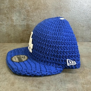 May include: A blue crochet baseball cap with a white logo on the front. The cap has a curved brim and a New Era logo on the side. The cap is a snapback style.