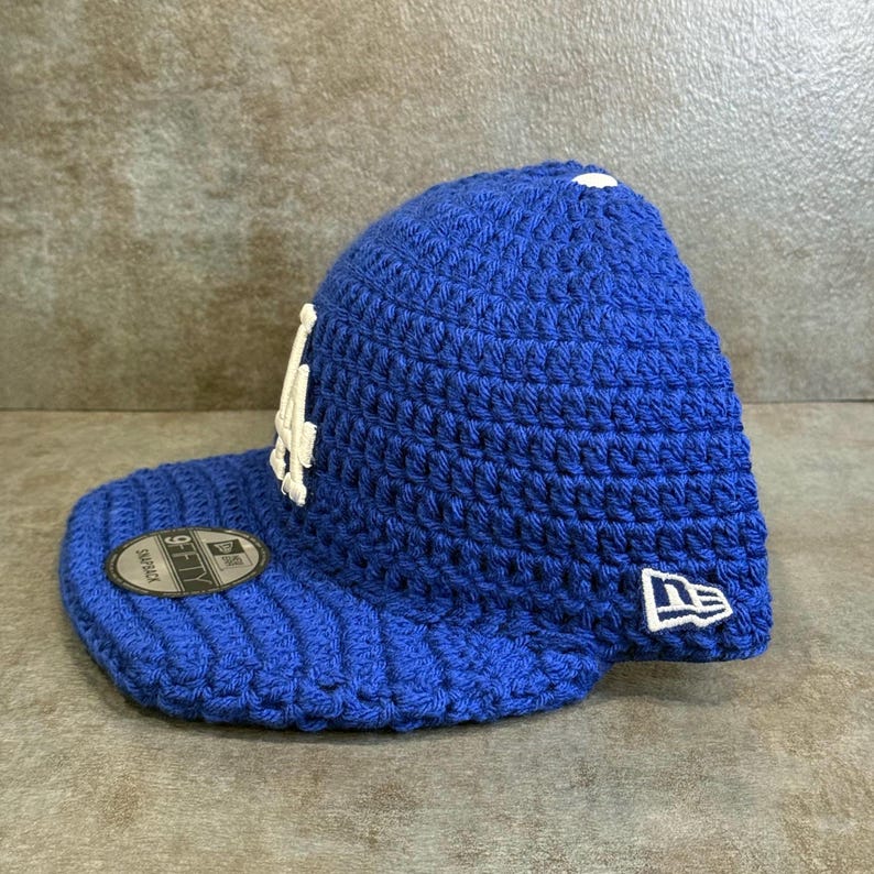 May include: A blue crochet baseball cap with a white logo on the front. The cap has a curved brim and a New Era logo on the side. The cap is a snapback style.