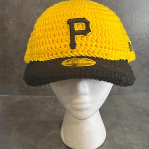 May include: A yellow and black crochet baseball cap with the letter "P" in black on the front. The cap has a black brim and is displayed on a white mannequin head. The cap is a sports-themed item.