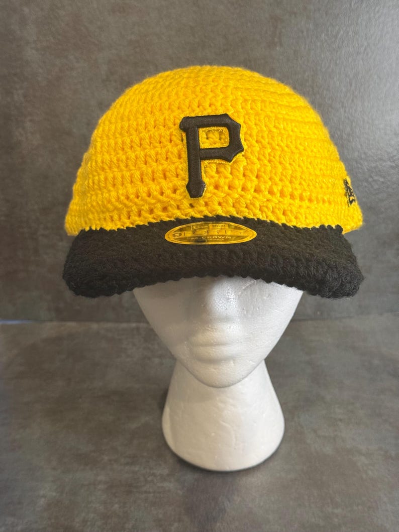 May include: A yellow and black crochet baseball cap with the letter "P" in black on the front. The cap has a black brim and is displayed on a white mannequin head. The cap is a sports-themed item.