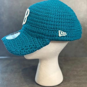 May include: Teal crochet baseball cap with a curved brim. The cap features a white "B" on the front and a white logo on the side. The cap is displayed on a white mannequin head.