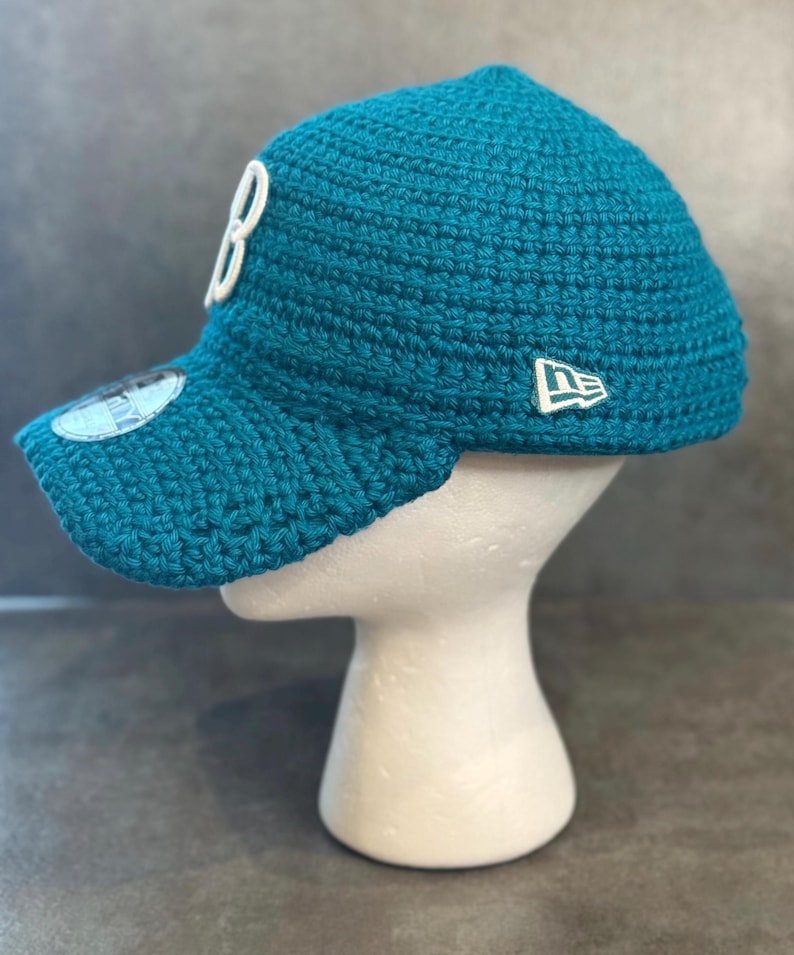 May include: Teal crochet baseball cap with a curved brim. The cap features a white "B" on the front and a white logo on the side. The cap is displayed on a white mannequin head.