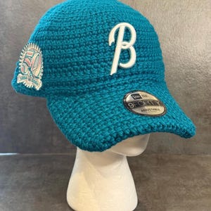 May include: Teal crochet baseball cap with a white "B" logo and a "50" patch. The cap has a curved brim and an adjustable strap. The "9FORTY" label is visible on the brim.
