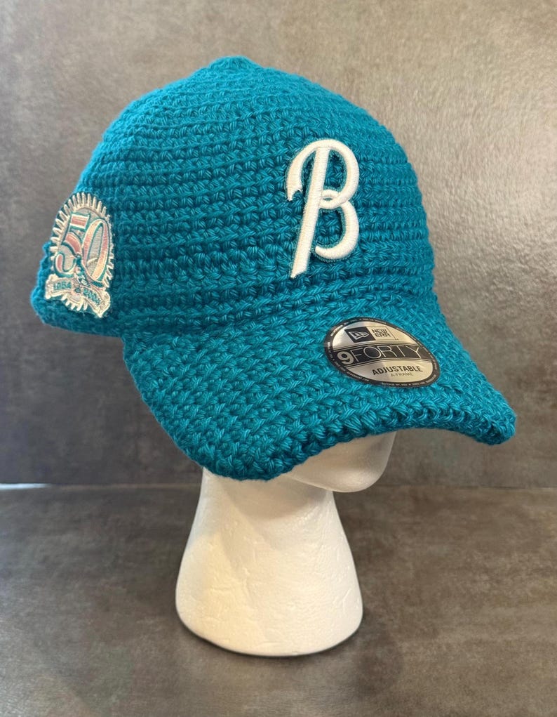 May include: Teal crochet baseball cap with a white "B" logo and a "50" patch. The cap has a curved brim and an adjustable strap. The "9FORTY" label is visible on the brim.
