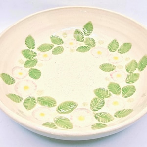 May include: A cream-coloured ceramic serving bowl with a floral design. The bowl features a ring of green leaves and white flowers with yellow centres. The rim of the bowl is slightly raised, and the interior has a speckled texture.