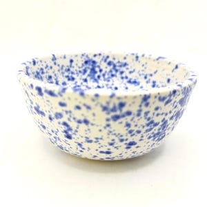 May include: A small, off-white ceramic bowl with a speckled blue pattern. The bowl has a rounded shape and a slightly flared rim. The blue speckles are irregularly sized and distributed across the surface.