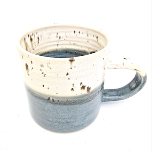 May include: A ceramic mug with a speckled, cream-coloured upper section and a blue-grey lower section. The mug has a rounded form and a robust handle. The interior is a darker shade of blue-grey. A unique piece for enjoying hot drinks.