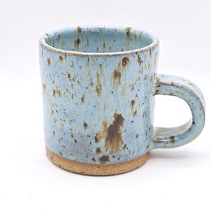 May include: A handmade ceramic mug with a light blue speckled glaze and a brown base. The mug has a rounded handle and a cylindrical shape. The mug is approximately 10 cm tall.