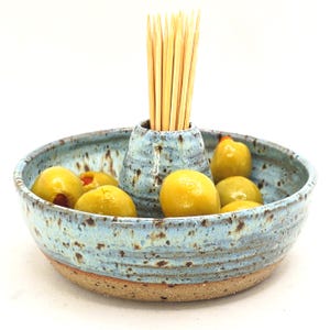 May include: A round ceramic serving bowl with a speckled light blue glaze and a natural tan base. The bowl features a central cone-shaped holder filled with wooden toothpicks. Several green olives are arranged in the basin around the centre.