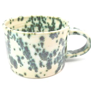 May include: A ceramic mug with a speckled glaze. The mug has a cream-coloured base with dark teal and grey spots. It has a rounded body, a handle, and a capacity of approximately 350 ml. The mug is suitable for hot or cold drinks.