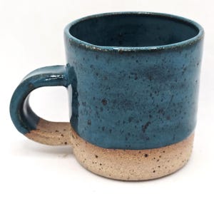 May include: A handmade ceramic mug. The upper half is glazed in teal blue, the base is natural speckled tan. It has a rounded handle and flared rim. The mug is approx. 10 cm tall. Ideal for tea or coffee.