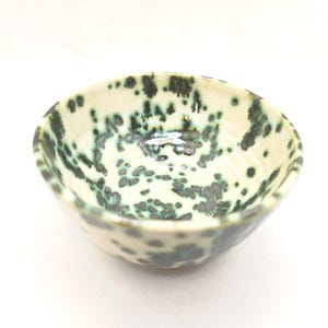 May include: A small ceramic bowl with a speckled pattern. The bowl has a creamy white base with dark green and grey spots. The interior is smooth and glossy, while the exterior has a slightly textured feel. The bowl is approximately 10 cm in diameter.